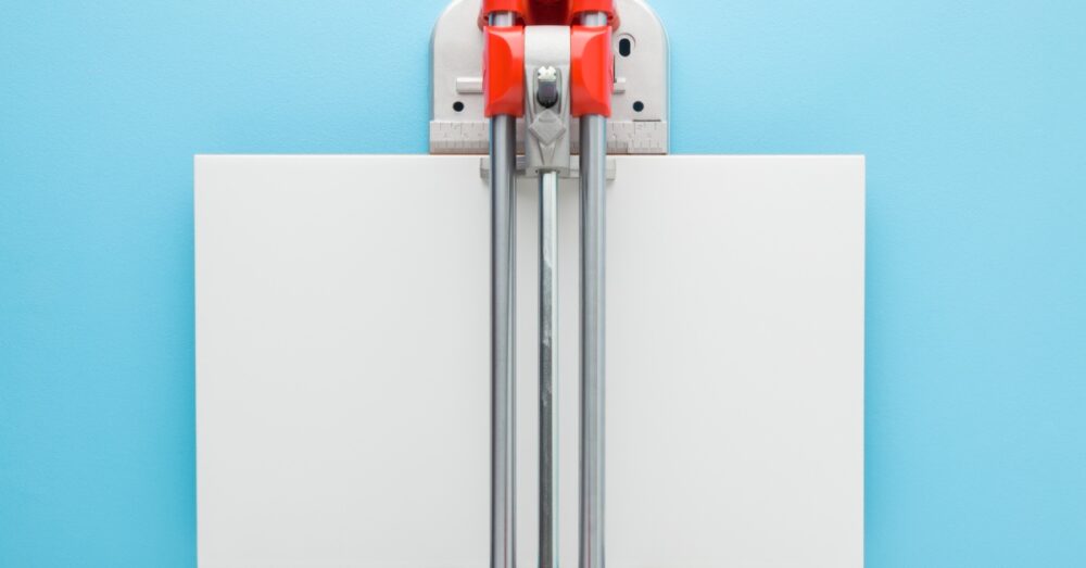 A red tile cutter loaded with a white ceramic tile set against a solid color, pastel blue background.