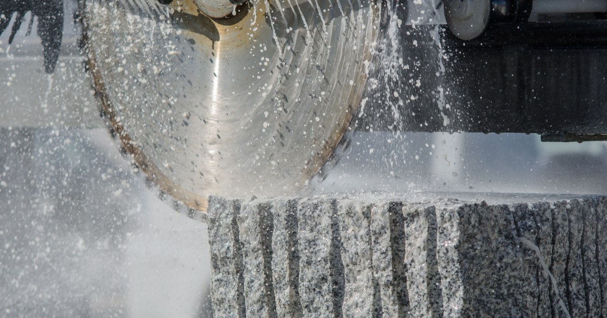 A close-up of the blade of a large industrial saw cutting into a block of grey and white granite stone.