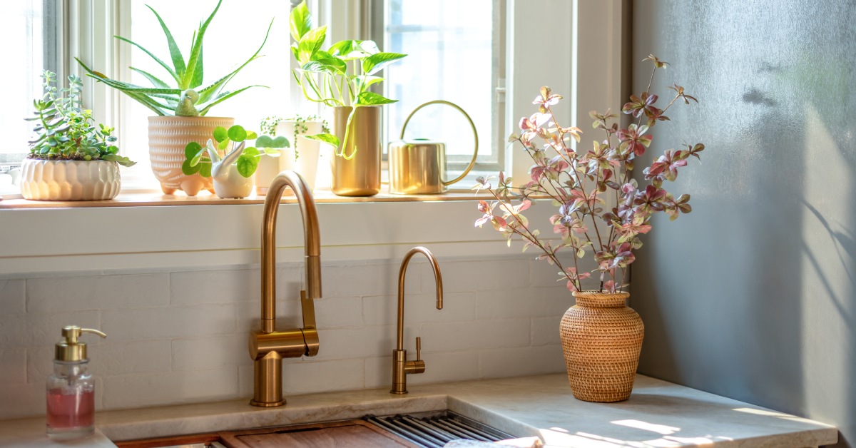 A home's quartzite kitchen counter with a sink and a window above it. The counter includes brass faucets and various flowers.