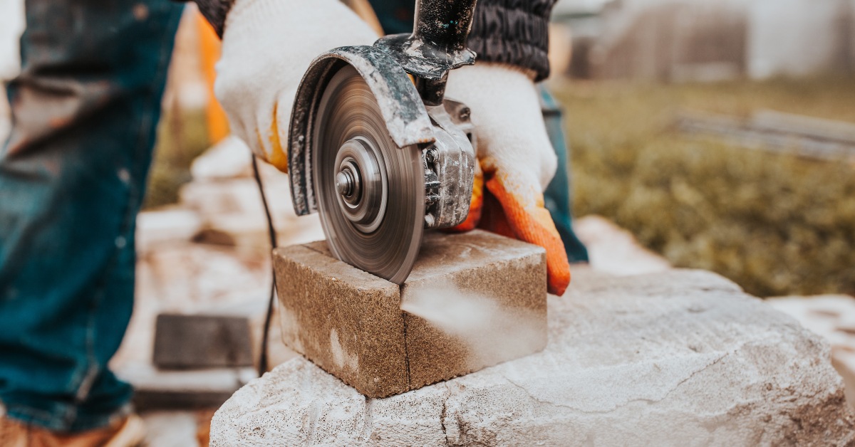 A man wearing protective working gloves holds a diamond cutter and saws into a brick slab atop a concrete block.