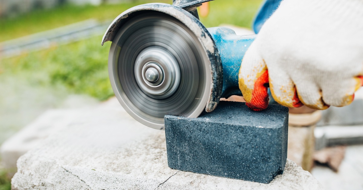 A person holds a sharp diamond cutting tool, sawing into a black stone atop a slab of concrete outside.