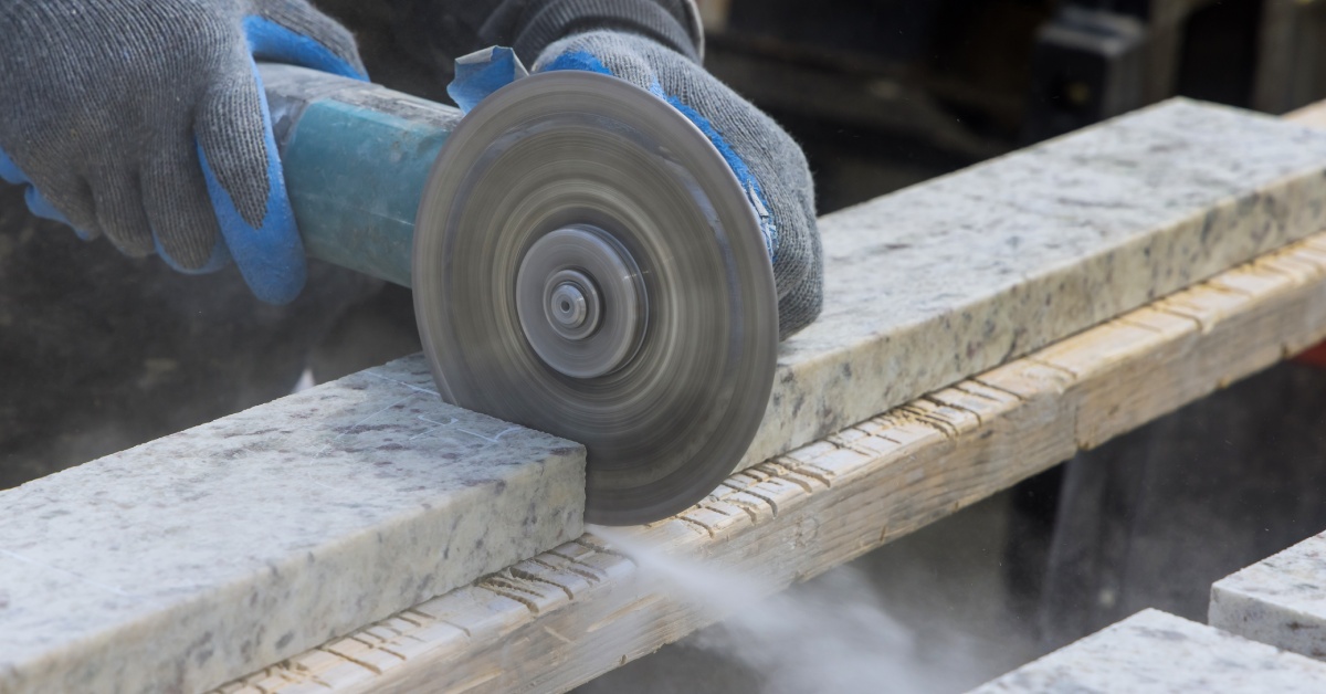 A person holds a diamond cutting tool with two hands while sawing into a granite plate. Dust blows out from the blade.