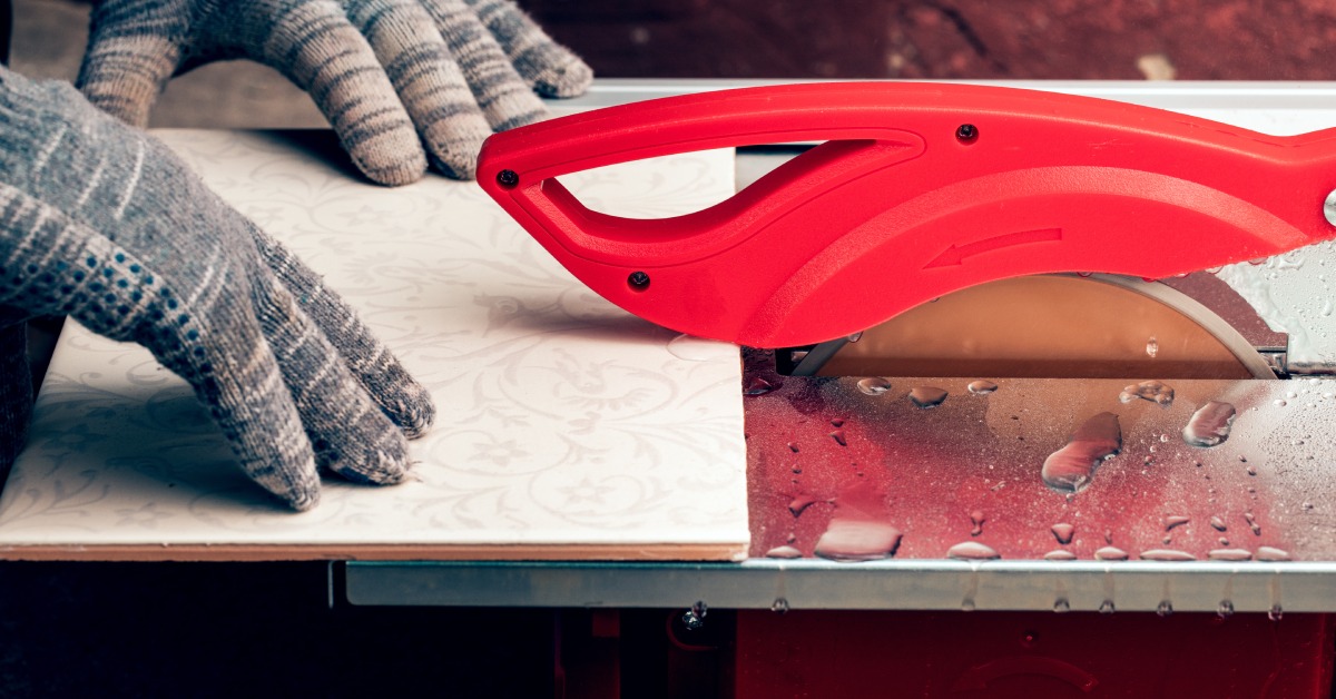 Gloved hands guide a patterned white tile through an electric tile cutter with a red handle and wet metal base.