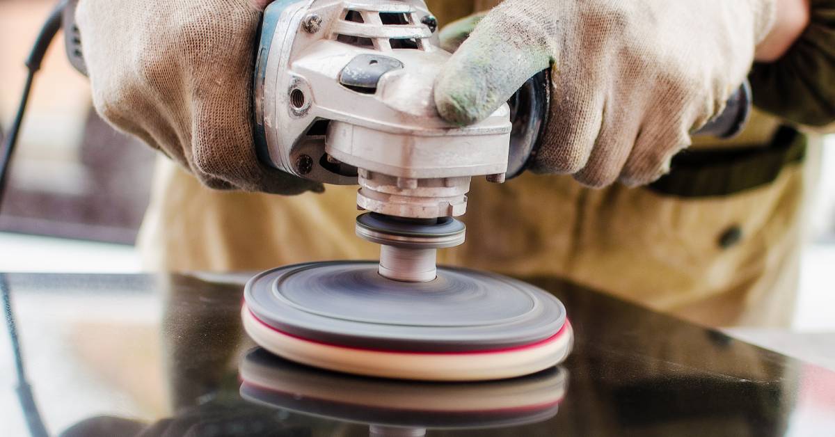 A close-up of a worker wearing gloves and an apron, holding an electronic polisher against a black stone countertop.