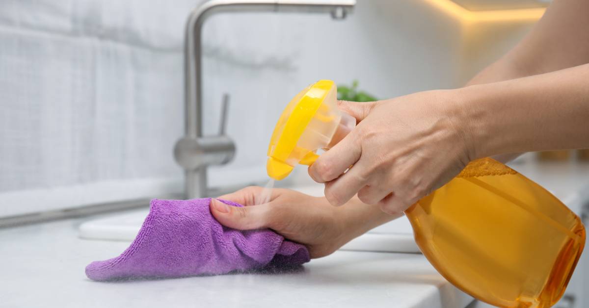 A woman's hands use a yellow spray bottle to spray cleaning solution onto a marble countertop. She uses with a purple cloth.