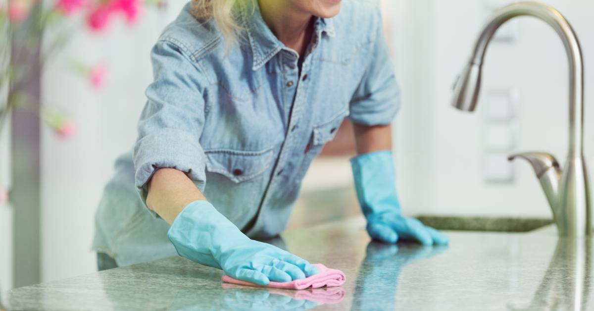 A woman wearing a denim top and blue latex gloves wipes the surface of marble kitchen countertops with a pink rag.