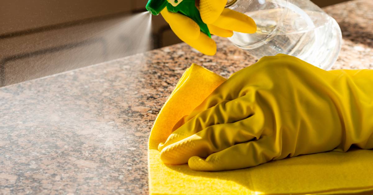 A pair of hands with yellow latex gloves spraying a countertop with a cleaning solution and wiping with a yellow rag.