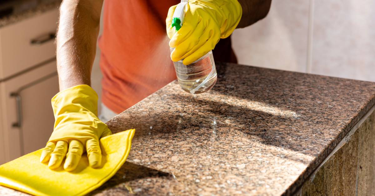 A man with latex gloves sprays a cleaning solution from a spray bottle onto a granite countertop and wipes with a yellow rag.