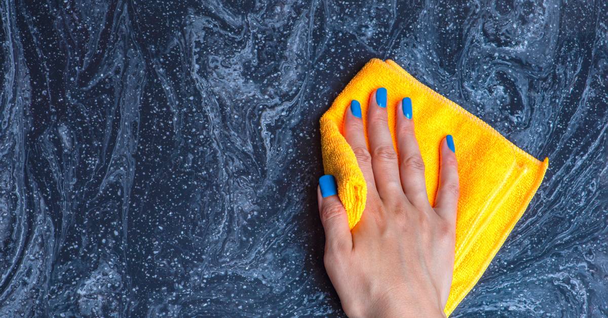 A close-up of a woman's hands with blue nail polish using a yellow rag to wipe at a blue-grey natural stone countertop.