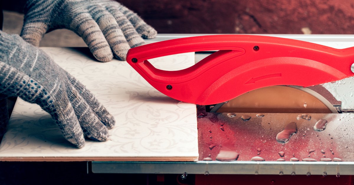 A worker with gloves slides a ceramic tile with decorative markings through an electric tile cutter saw.