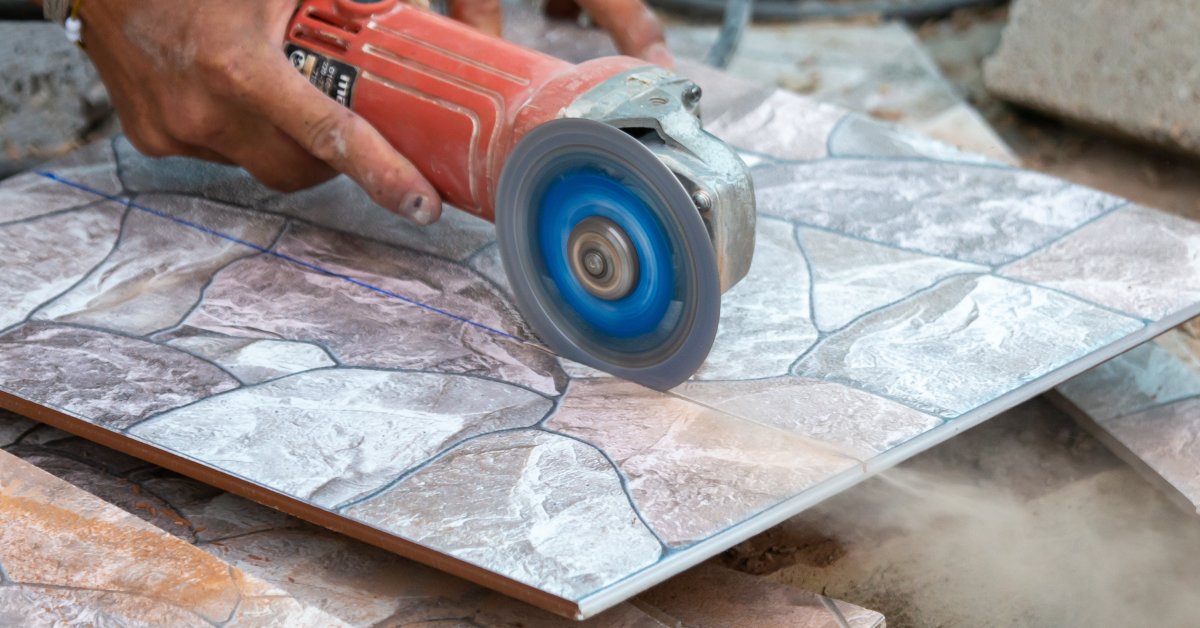 A close-up of a hand using an angle tile grinder to cut into a thin slab of granite floor tiling and creating dust.