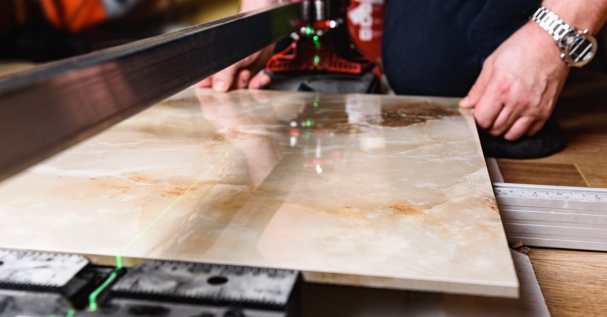 A close-up of a man using a manual tile cutter with a green laser indicator to cut into a ceramic stone slab.