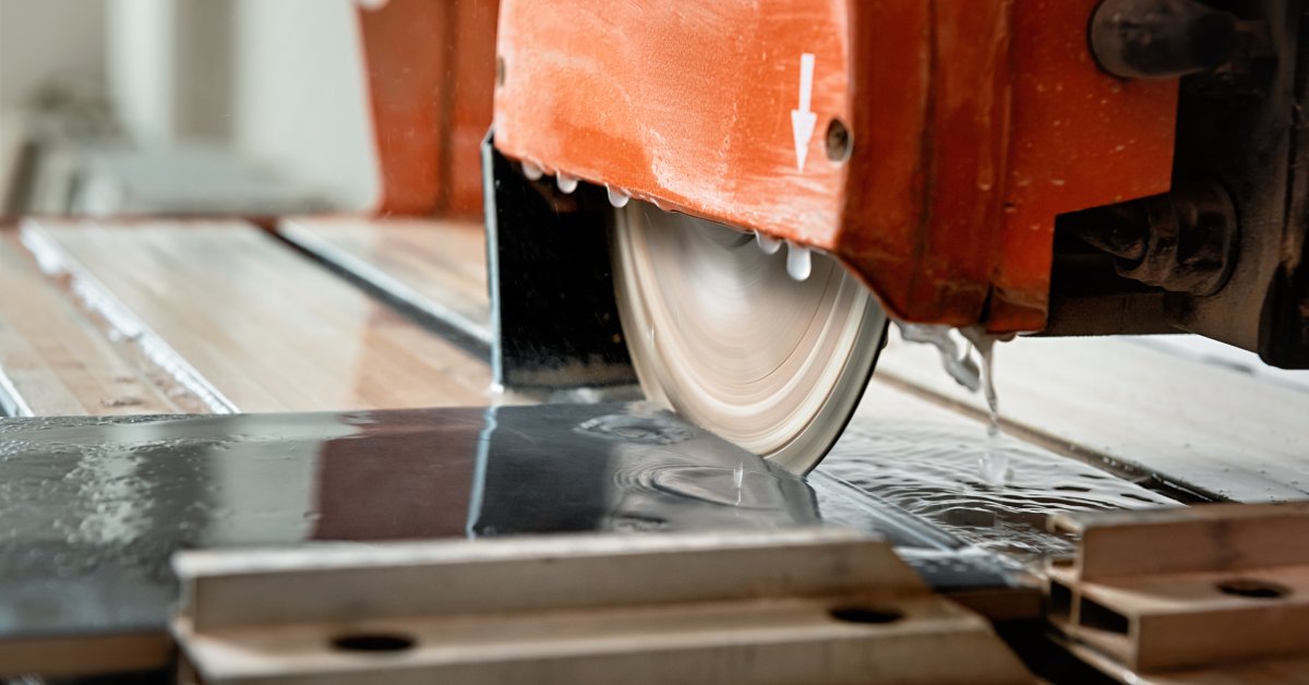 A wet diamond saw blade in a red cutting machine is cutting into a dark-grey slab of stone on a work bench.