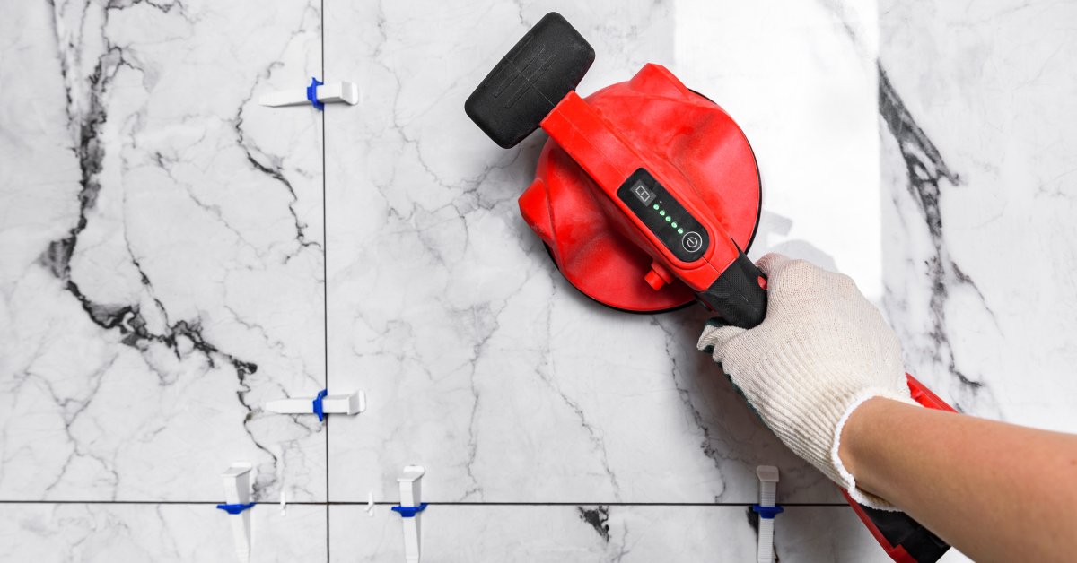 A close-up of a person's hand holding a red electric vibrating suction cup for tile handling on a white marble tile.