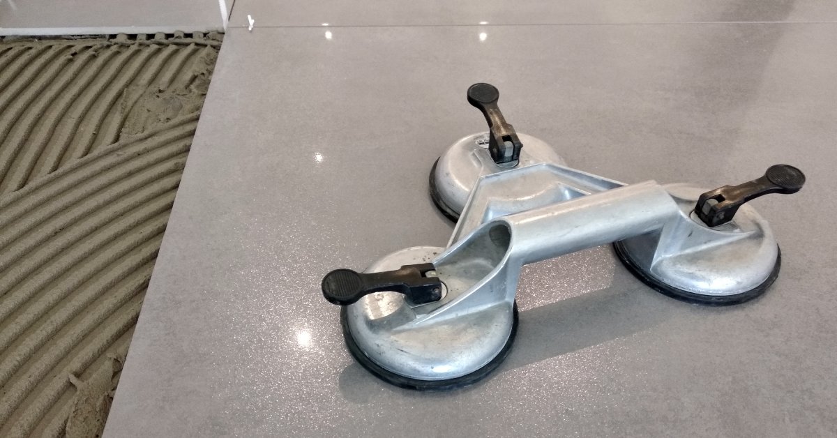 A close-up of a three-pronged suction cup device for moving tiles. The device is planted on a large grey floor tile.