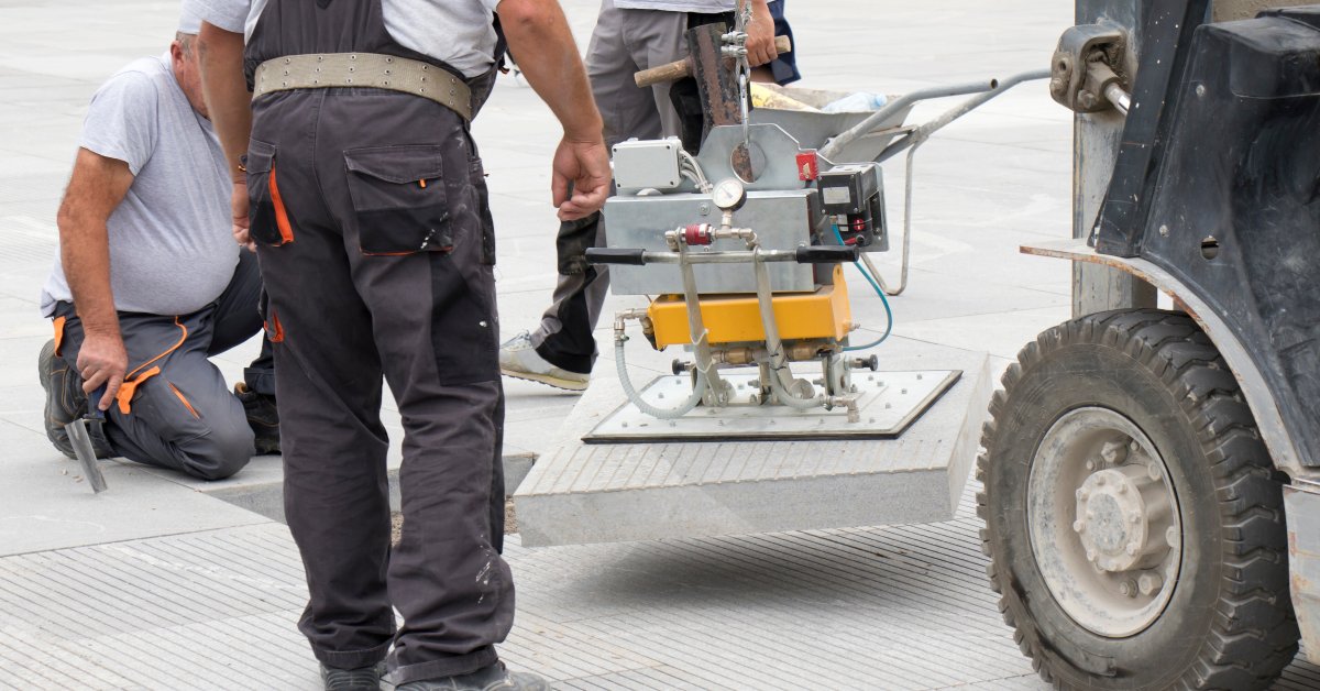 A group of three workers use a magnetic vacuum device to lift a large square block of pavement into an open space.