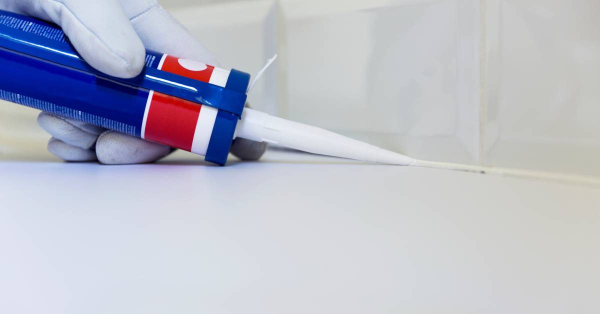 A close-up of a white-gloved hand holding a blue tube sealant applier and applying silicone sealant to a kitchen counter.