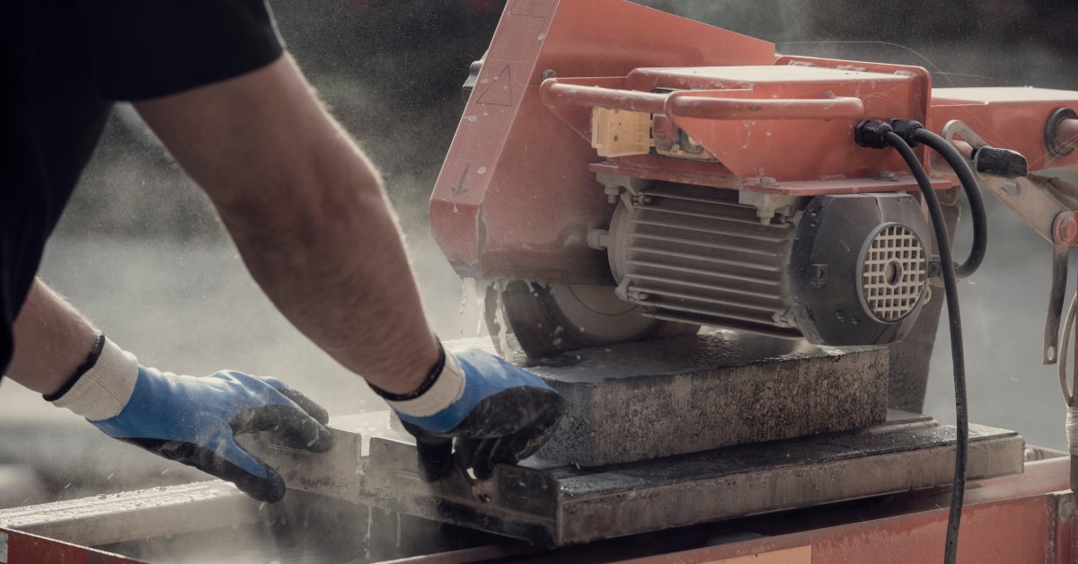 A worker with blue safety gloves feeds a slab of stone into a bridge saw machine. The saw sprays water as it cuts.