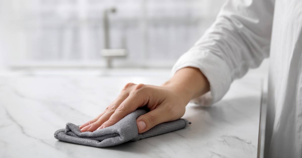 A close-up of a woman's hand wiping a white marble kitchen countertop in a white kitchen with a folded gray wash rag.