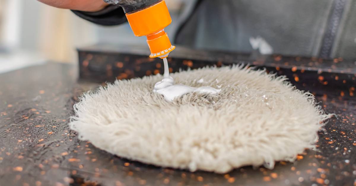 A close-up of a person applying a silver liquid polishing paste to a white polishing pad on a stone countertop.