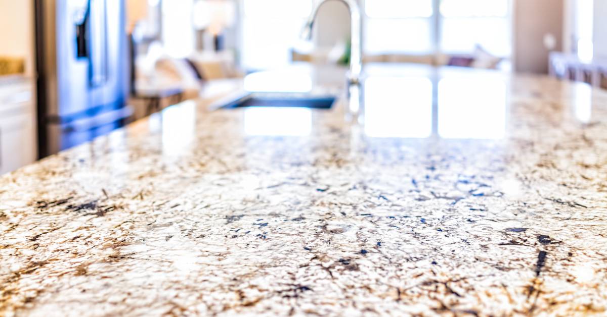 A close-up view of a granite kitchen island countertop that is white, brown, and black. The rest of the kitchen is blurred.