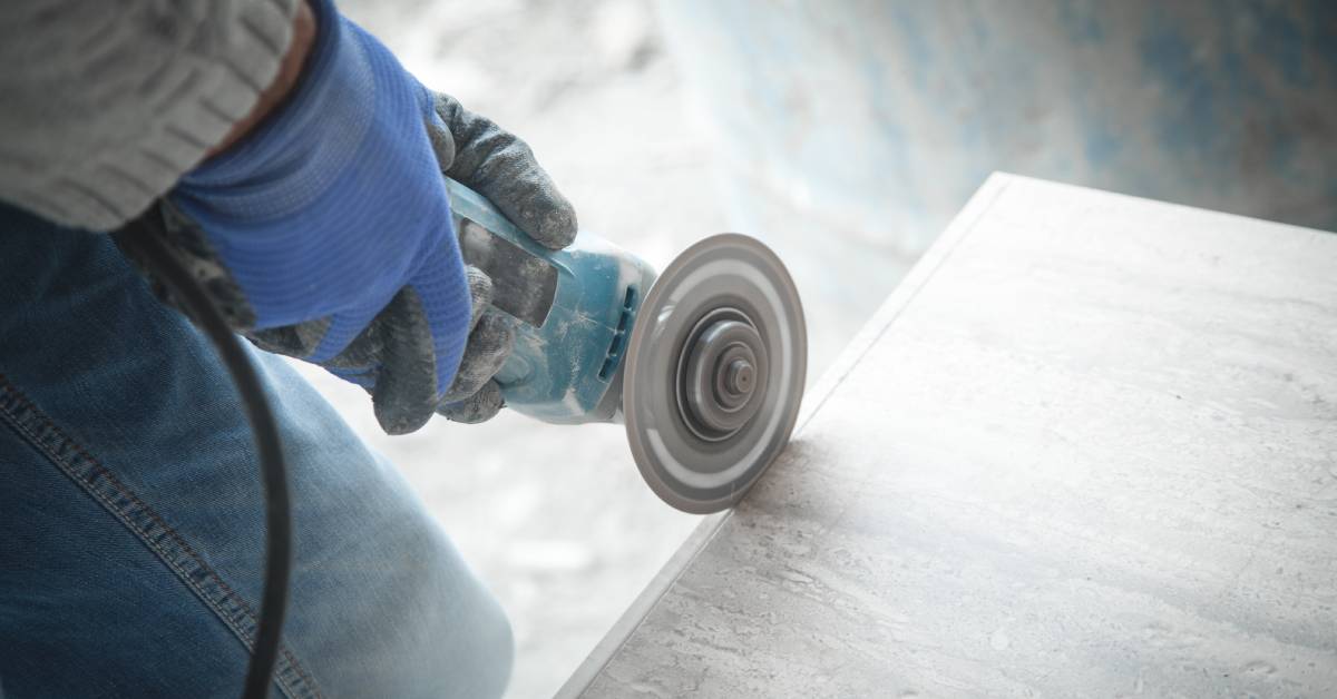 A close-up of a worker wearing jeans and gloves using an angle grinder to cut into a large square slab of white tile.