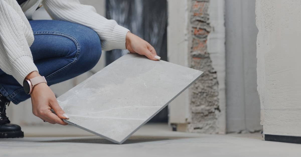 A woman in jeans and a white sweater crouches down to hold and examine a square porcelain floor tile.