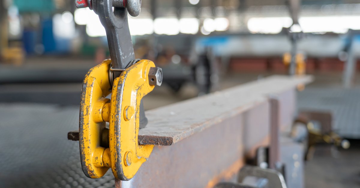 A close-up of a yellow steel clamp attached to an overhead crane holding onto a steel H-beam in a factory.