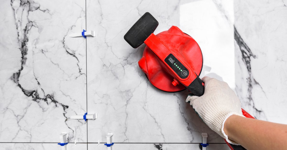 A close-up of a hand with a white glove holding a red vacuum suction cup lifter to a white tile square.