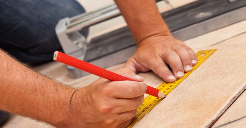 A close-up of a man kneeling and marking tile to be cut. The measuring tape is yellow and the tile is beige and brown.